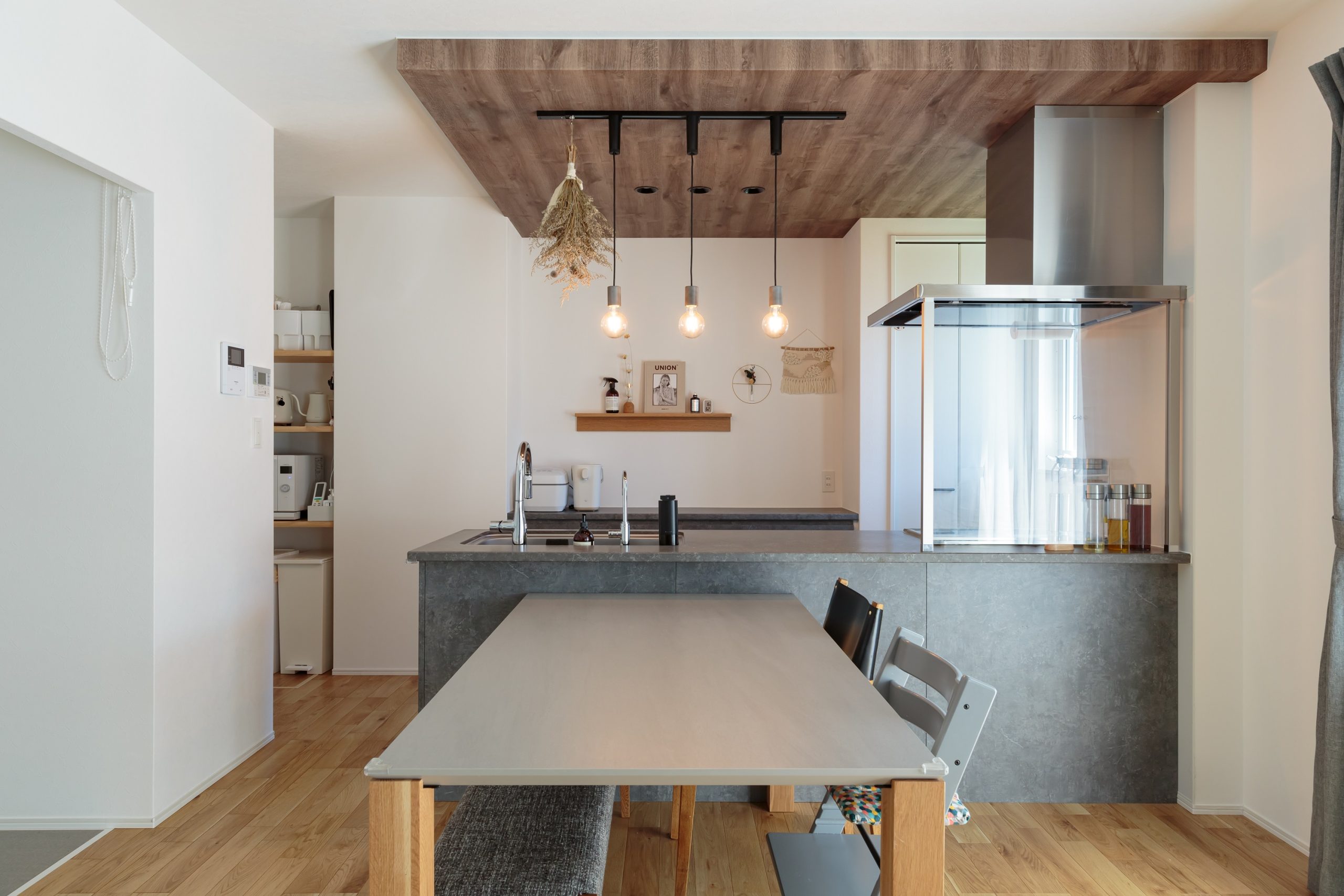 モルタル風キッチンが映えるようキッチン背面は白い壁ですっきりと、木目の下がり天井をアクセントに加えました。ダイニングテーブルもキッチンに質感を合わせてトータルコーディネートしています。